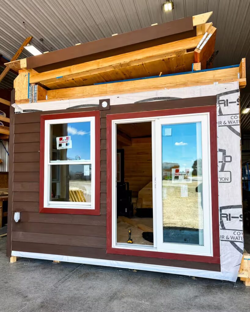 Prefab tiny house under construction with brown siding, red-trimmed window, and sliding glass door; roof framing and lumber stacked on top.
